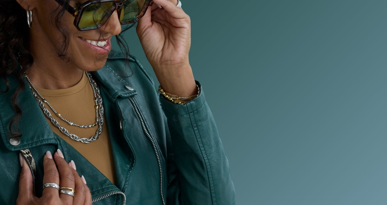 Frau mit Sonnenbrille und grünem Lederlook, die auffällige Ketten, Armreifen und Ringe in Gold und Silber aus der Jewels-Kollektion trägt.