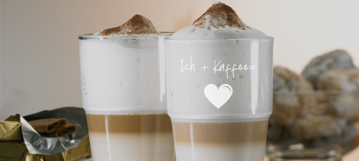 Zwei gravierte Latte-Macchiato-Gläser von LEONARDO mit der Aufschrift „Ich + Kaffee = ❤️“ gefüllt mit Milchschaum und Schokopulver.