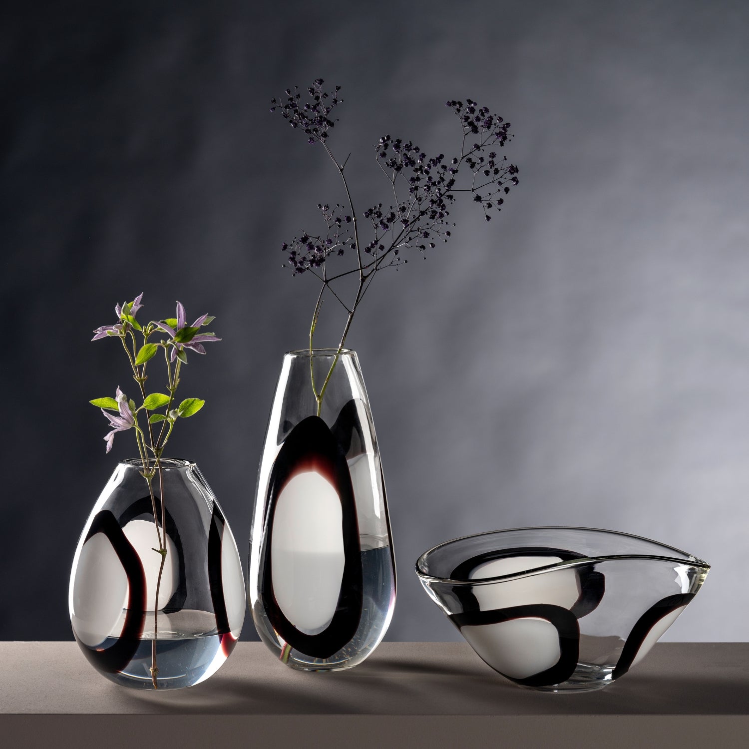 Zwei mundgeblasene Vasen und eine passende Schale aus klarem Glas mit kunstvollem schwarz-weißem Dekor stehen vor dunklem Hintergrund und setzen einzelne Zweige stilvoll in Szene.