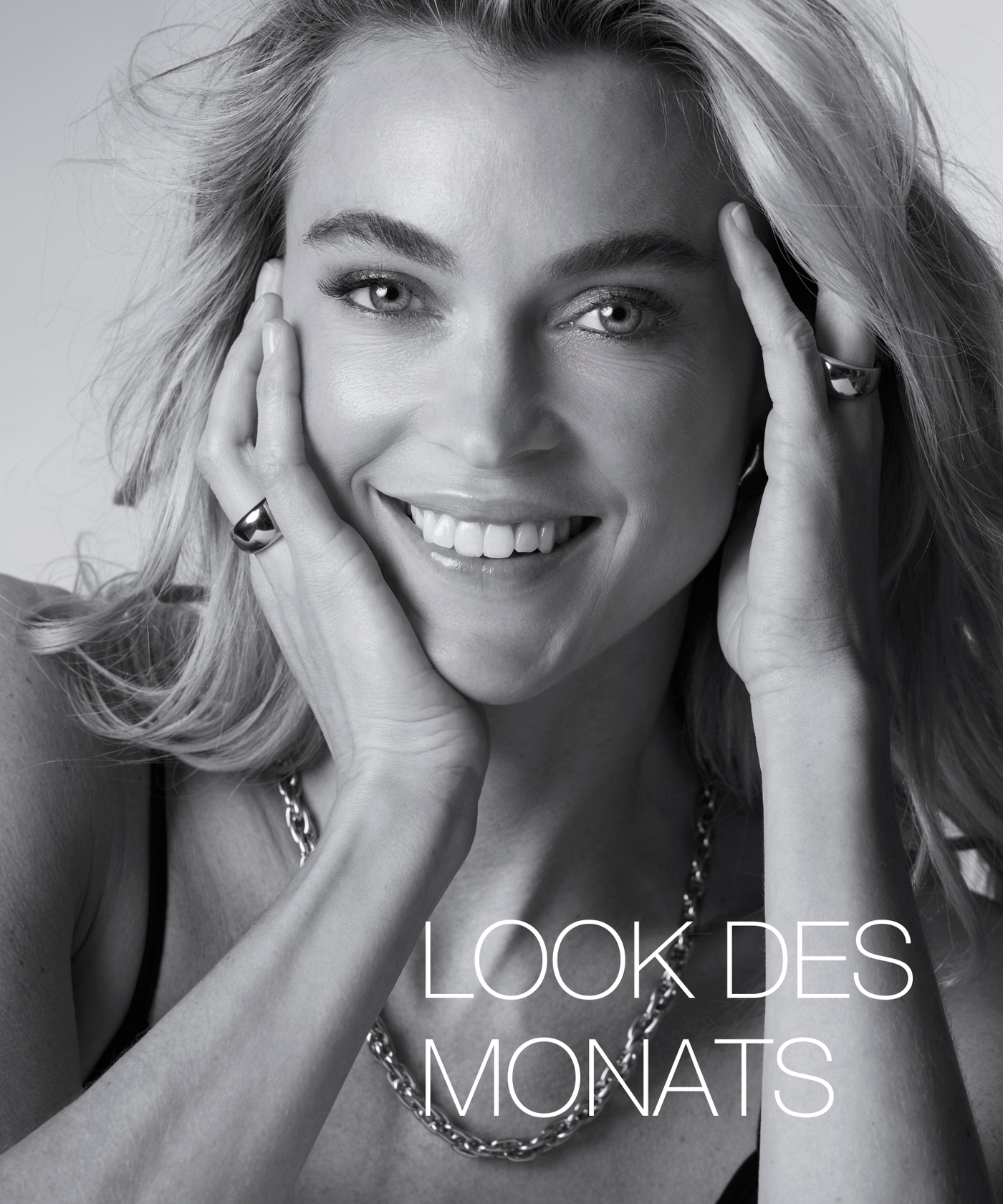 Schwarz-weiß-Porträt einer lächelnden Frau mit offenen Haaren, die ihr Gesicht mit beiden Händen berührt und eine Kette sowie Ringe trägt. Schriftzug ‚Look des Monats‘ im Bild.