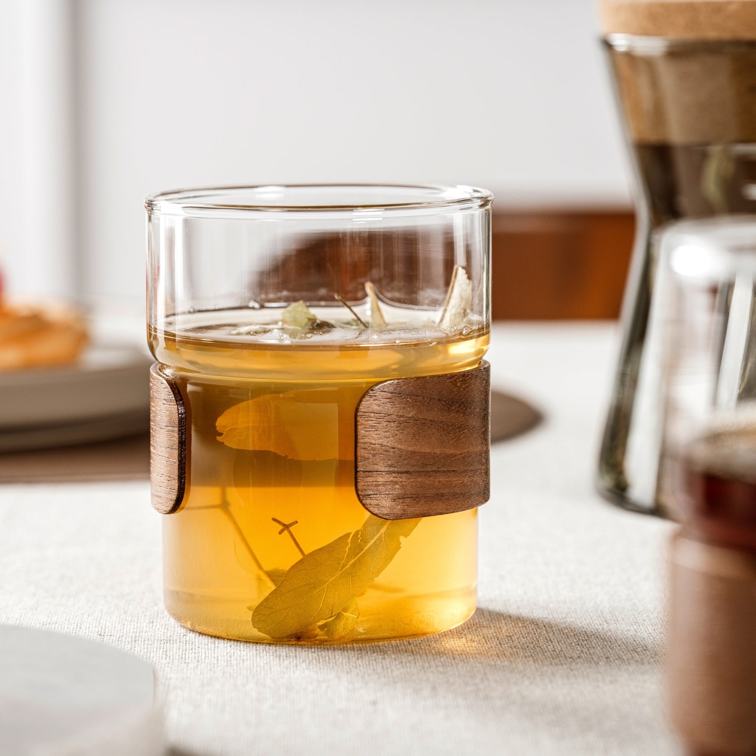 Ein stilvolles Teeglas mit dekorativer Holzmanschette zeigt einen frisch aufgebrühten Kräutertee mit sichtbaren Blättern und fügt sich harmonisch in das natürliche Tischambiente ein.