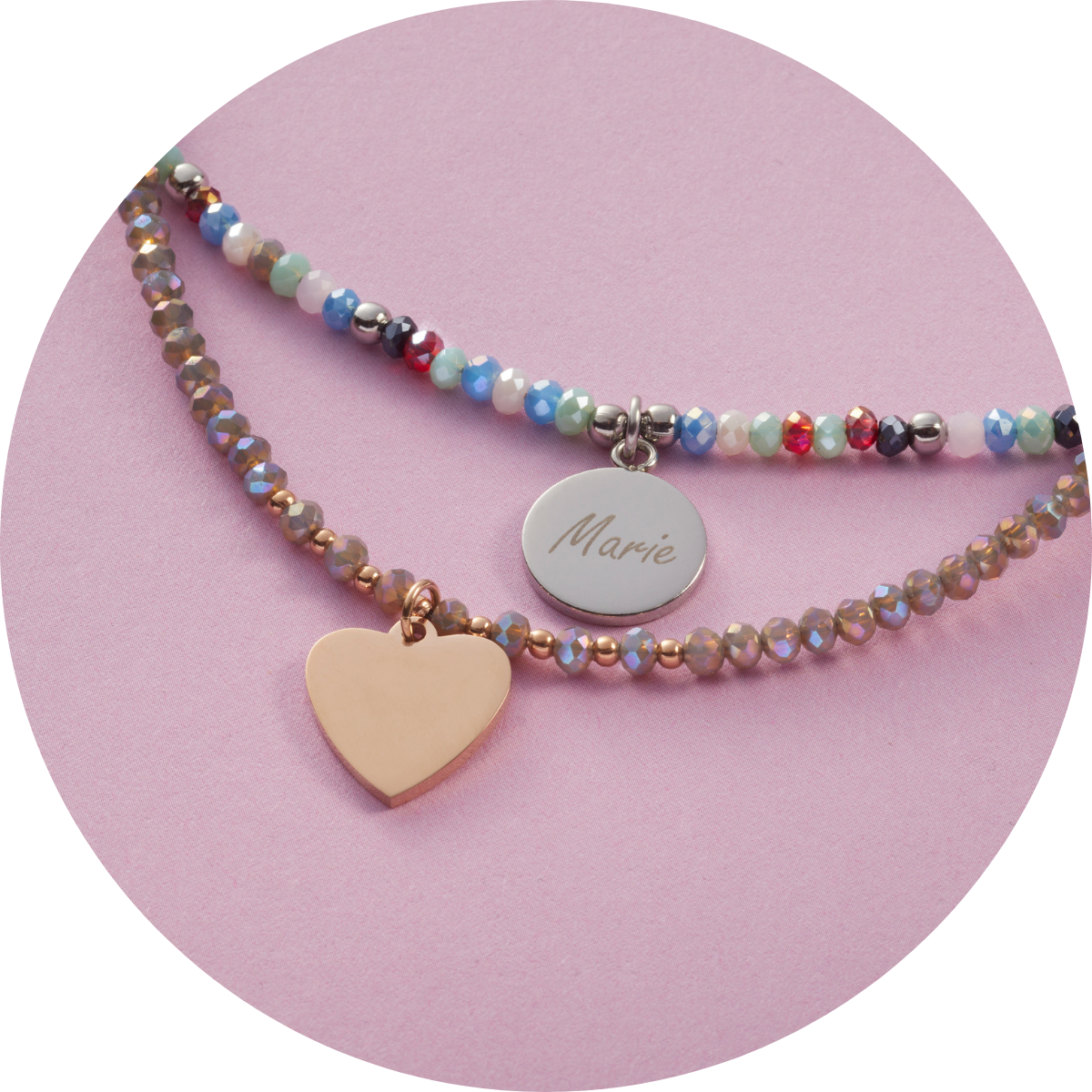 Zwei bunte Armbänder auf rosafarbenem Untergrund – eines mit facettierten Perlen und gravierter Herz-Plakette in Roségold, das andere mit bunten Glasperlen und rundem Edelstahl-Anhänger mit dem Namen „Marie“.