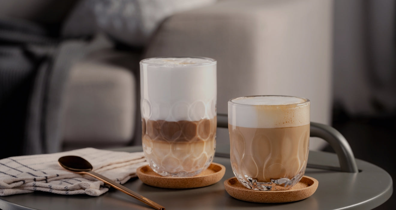 Zwei Cappuccinos mit Milchschaum in strukturierten Gläsern auf Korkuntersetzern, serviert auf einem Tablett im gemütlichen Wohnzimmerambiente.