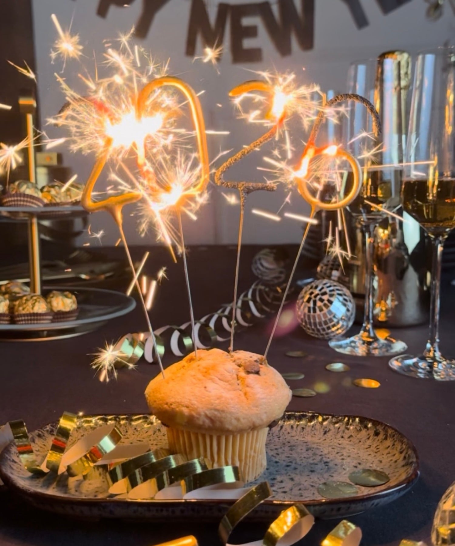 Silvesterdekoration mit einem Muffin auf einem Teller, in den brennende Wunderkerzen in Form der Jahreszahl 2026 gesteckt sind; im Hintergrund stehen Sektgläser, goldene Konfetti und Partydeko.