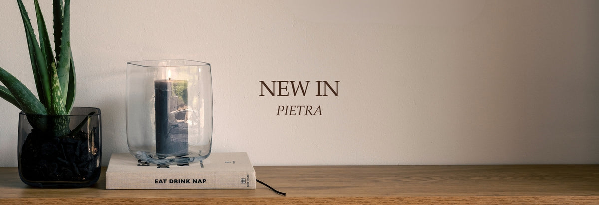 NEW IN PIETRA: Klares Glas mit Wasser und Blausteinen neben Sukkulente, Buch und heller Holzoberfläche
