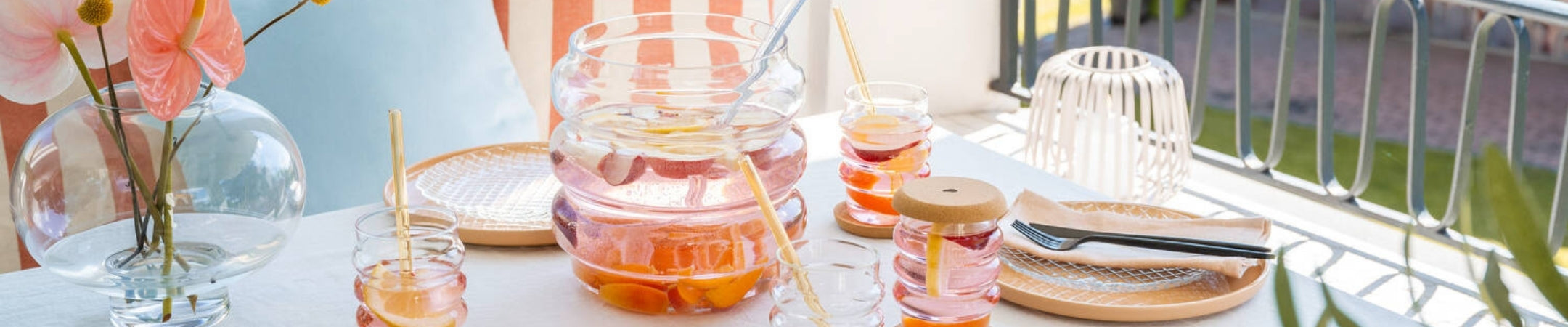Sommerlich gedeckter Tisch auf dem Balkon mit der geschwungenen SOLE-Bowle aus Glas, passenden Trinkgläsern mit Obstbowle, Glastrinkhalmen und farblich abgestimmter Dekoration in Rosa, Apricot und Naturtönen.