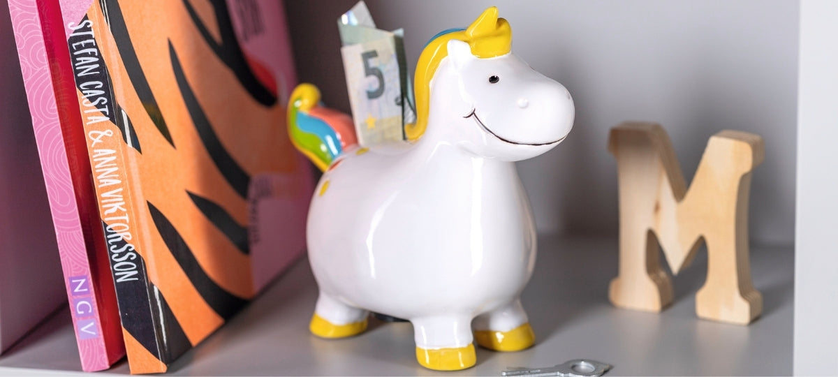 Weiße Spardose in Unicorn-Form mit goldenen Details, umgeben von Büchern und Holzbuchstaben auf grauem Regal