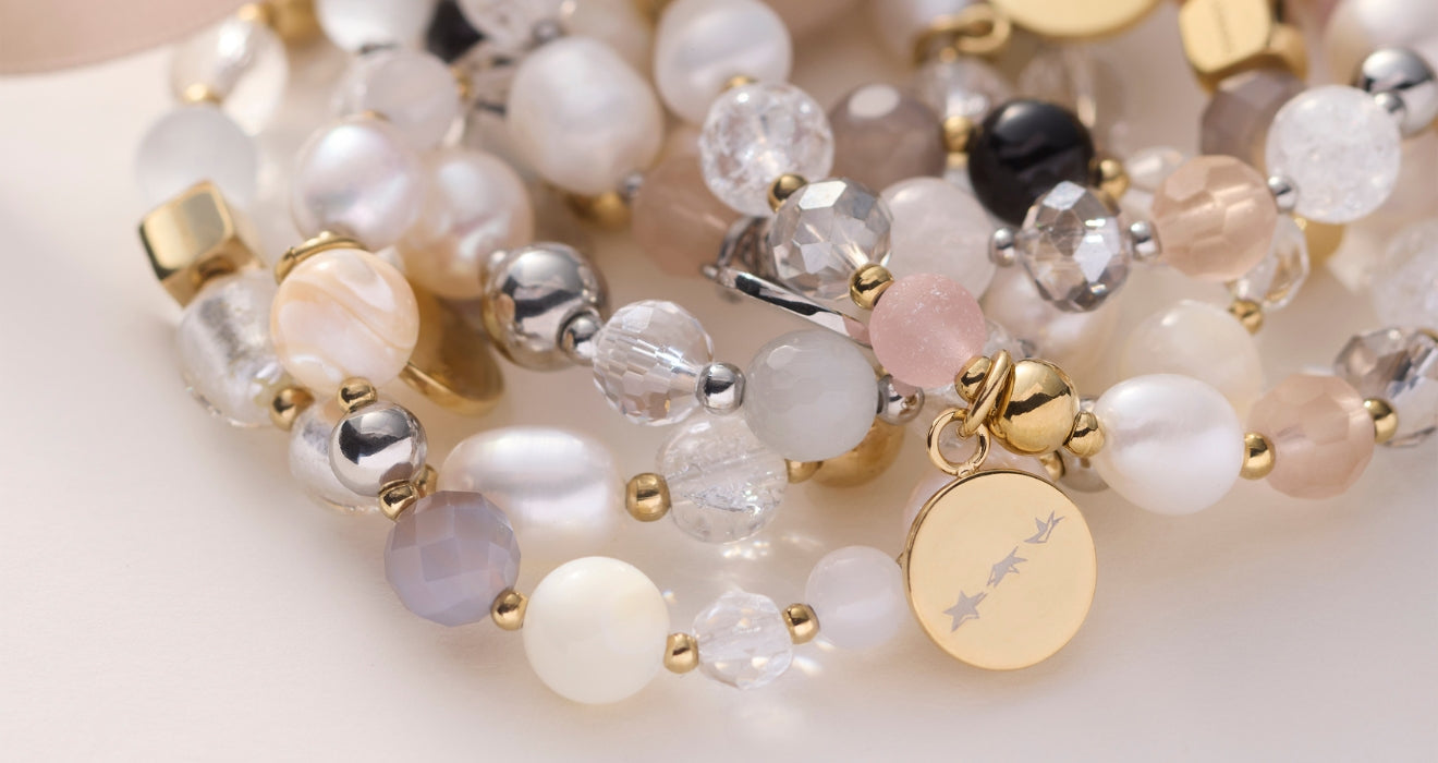 Ein Armband aus verschiedenfarbigen Perlen und Edelsteinen in zarten Pastelltönen liegt kunstvoll arrangiert – ein kleiner runder Anhänger mit Sternen rundet das Design liebevoll ab.