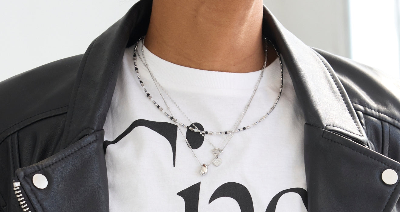 Zwei filigrane Halsketten mit silbernen und schwarzen Perlen sowie kleinen Anhängern werden lässig über einem weißen T-Shirt mit Lederjacke getragen.