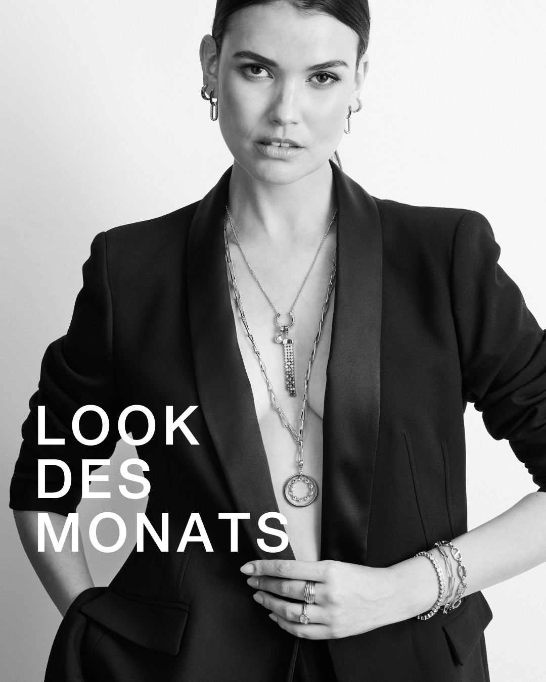 Frau in elegantem schwarzen Blazer mit mehreren silberfarbenen Halsketten und auffälligem Armschmuck. Der moderne Look kombiniert zeitlose Eleganz mit starkem Ausdruck. Text: LOOK DES MONATS.