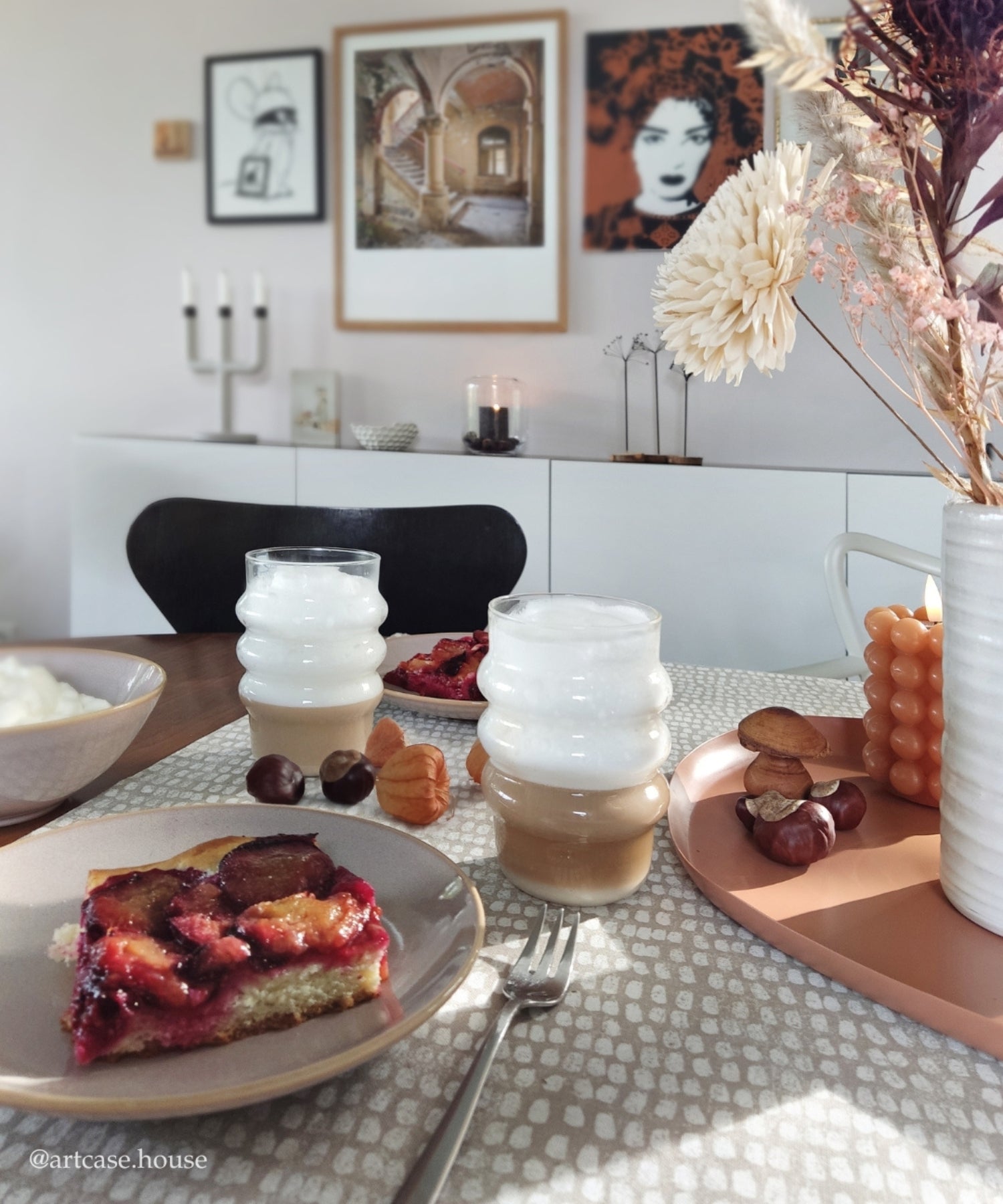 Gemütlich gedeckter Tisch mit Latte Macchiato in organisch geformten Gläsern, Zwetschgenkuchen und herbstlicher Deko aus Kastanien und Trockenblumen.