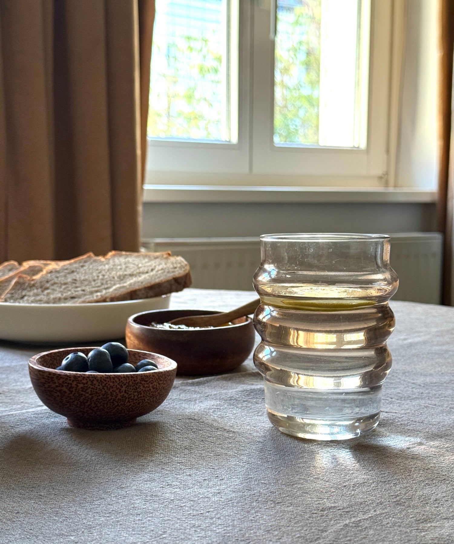 Klares Wasserglas mit wellenförmiger Struktur auf einem Frühstückstisch mit Brot, Marmelade und frischen Blaubeeren in Schälchen.