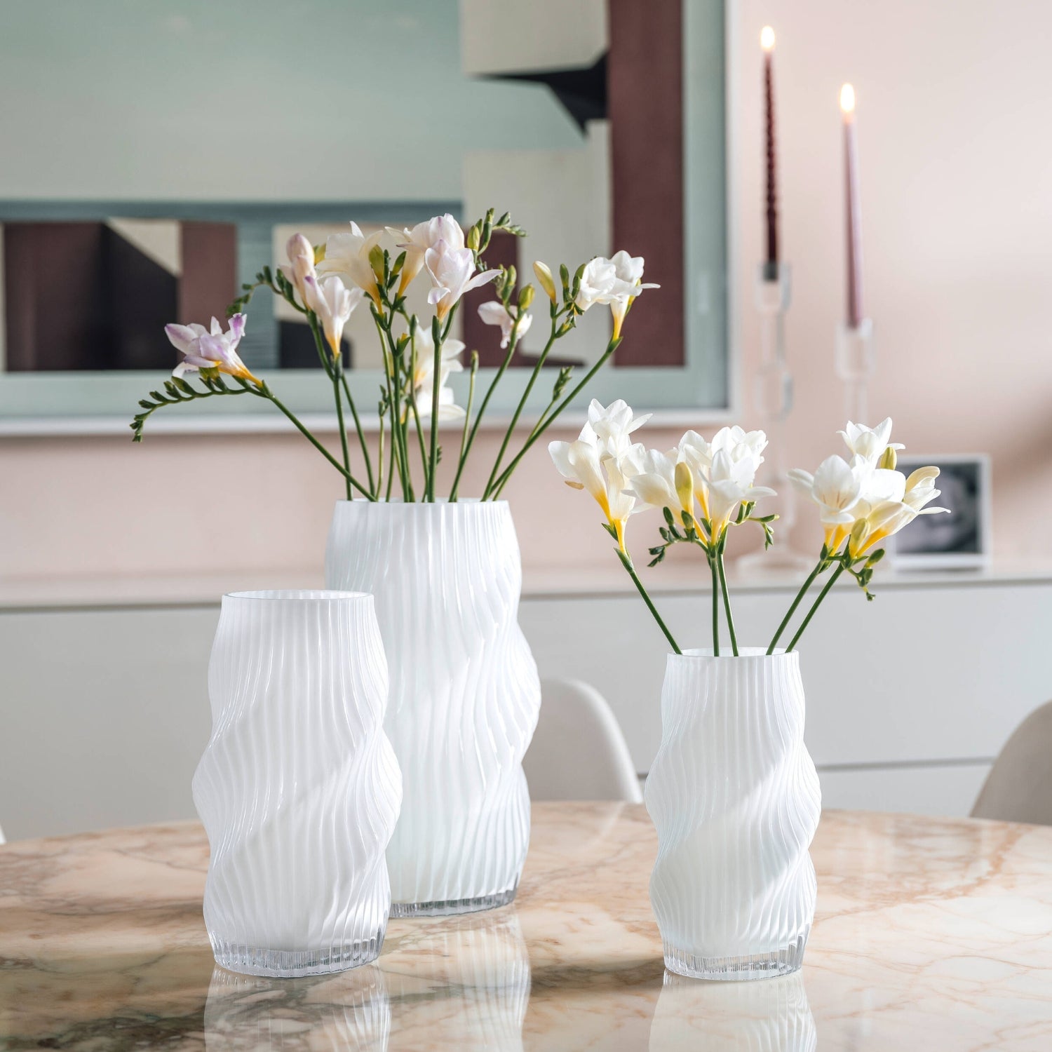 Drei weiße Glasvasen mit organisch geschwungenem Rillenmuster sind auf einem runden Marmortisch dekorativ mit zarten Freesienblüten arrangiert.