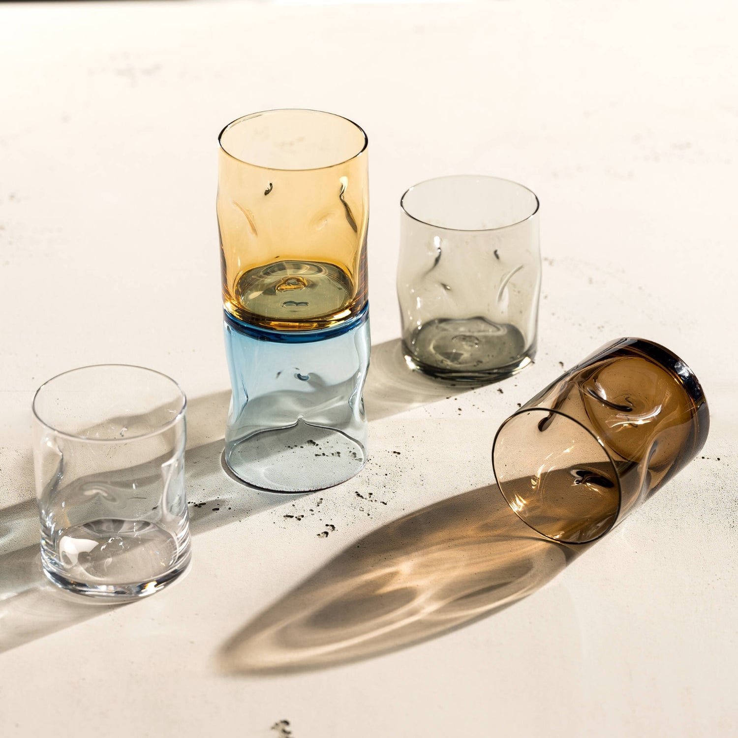 Fünf farbige Trinkgläser mit markanten Einkerbungen liegen und stehen locker arrangiert auf einem hellen Untergrund und werfen lebendige, organische Schatten – jedes Glas ein individuelles Designobjekt.