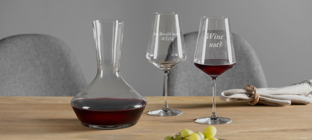 Zwei Weingläser von LEONARDO mit den Sprüchen „Im Zweifel mehr WEIN“ und „Wine not?“ neben einer eleganten Karaffe auf gedecktem Tisch.