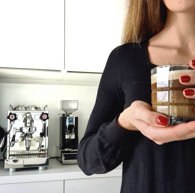 Frau in schwarzem Oberteil hält ein Glas Latte Macchiato in der Hand, im Hintergrund moderne Siebträgermaschine und Kaffeemühle in heller Küche.