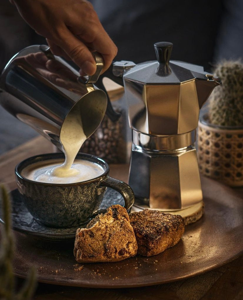 Szene mit einer Hand, die aufgeschäumte Milch in eine Tasse gießt, daneben eine klassische Espressokanne und zwei Stück Gebäck auf einem Tablett im gemütlichen Ambiente.