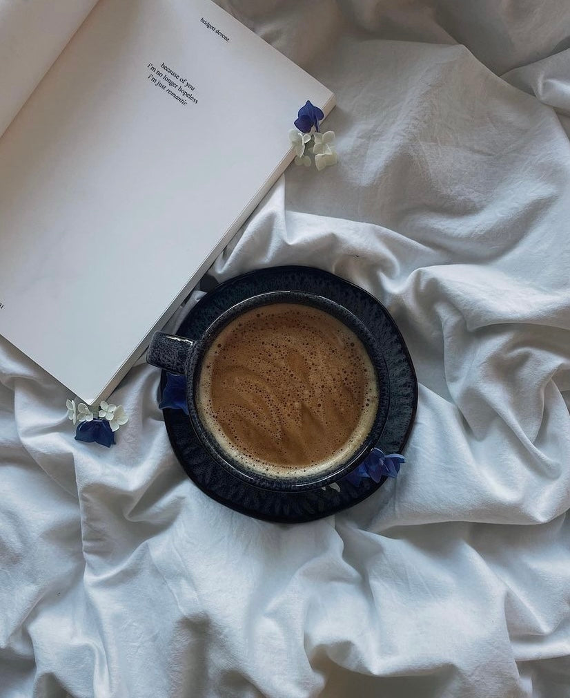 Dunkle Kaffeetasse auf einem Unterteller auf weißer Bettdecke, daneben ein aufgeschlagenes Buch und kleine blaue Blüten als dekoratives Detail.