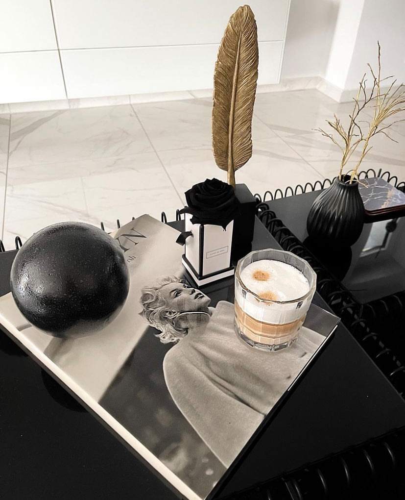 Flatlay mit einem Glas Cappuccino auf einem Bildband, dekoriert mit schwarzer Kugel, Duftflakon, goldener Feder und schwarzen Wohnaccessoires auf einem modernen Couchtisch.