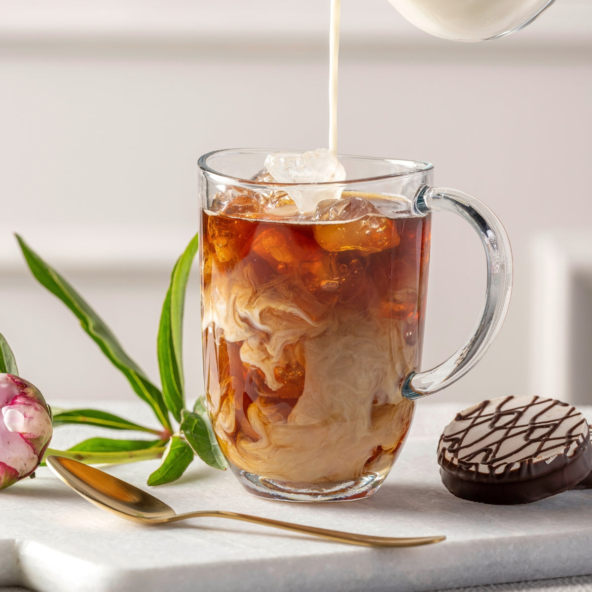 Ein Eiskaffee mit Milchwirbel in einer Glastasse, serviert mit Gebäck und goldfarbenem Löffel.