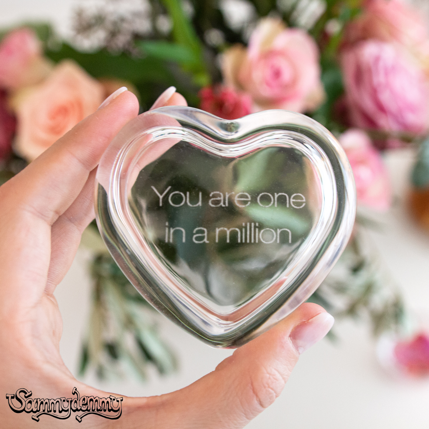 Eine Hand hält ein gläsernes Herz mit der eingravierten Botschaft „You are one in a million“, im Hintergrund ein bunter Strauß mit Rosen.