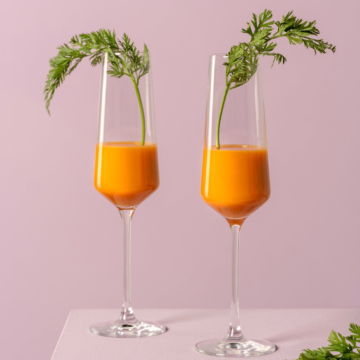Zwei elegante Sektgläser von LEONARDO, gefüllt mit leuchtend orangefarbenem Karottensaft und dekoriert mit frischem Karottengrün, vor einem pastellrosa Hintergrund.