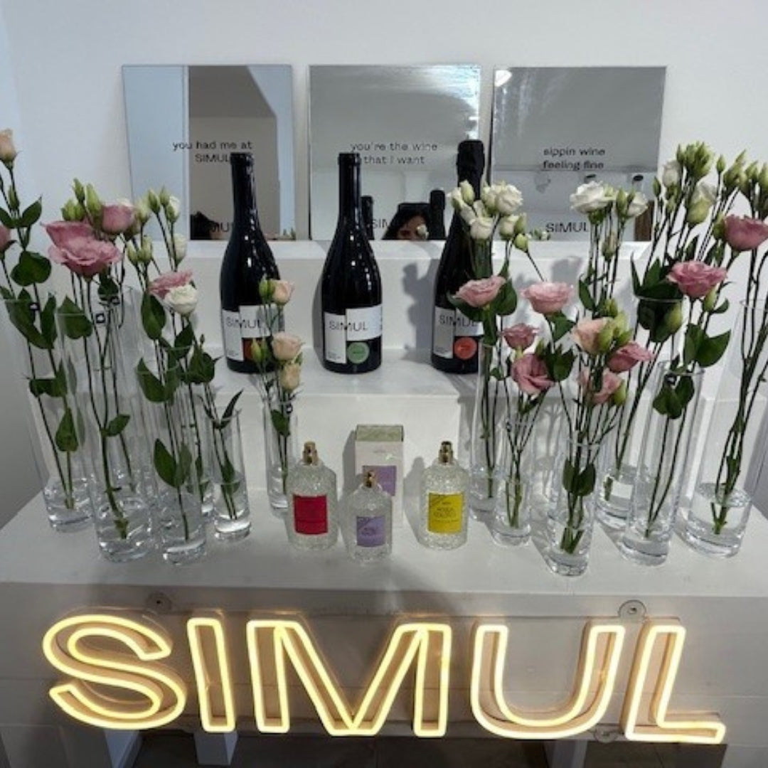 Elegante Produktpräsentation von SIMUL mit drei Weinflaschen, dekorativen Glasvasen mit rosa und weißen Blumen sowie Parfumflakons, stilvoll arrangiert auf einem Tisch mit leuchtendem SIMUL-Schriftzug.