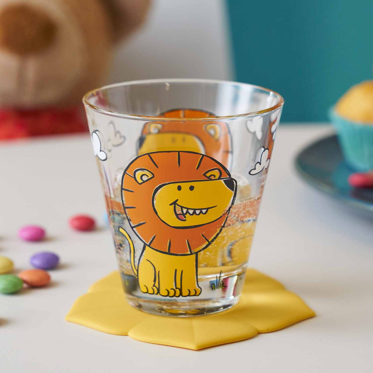 Trinkglas BAMBINI mit Löwenmotiv auf gelbem Untersetzer, umgeben von Bonbons