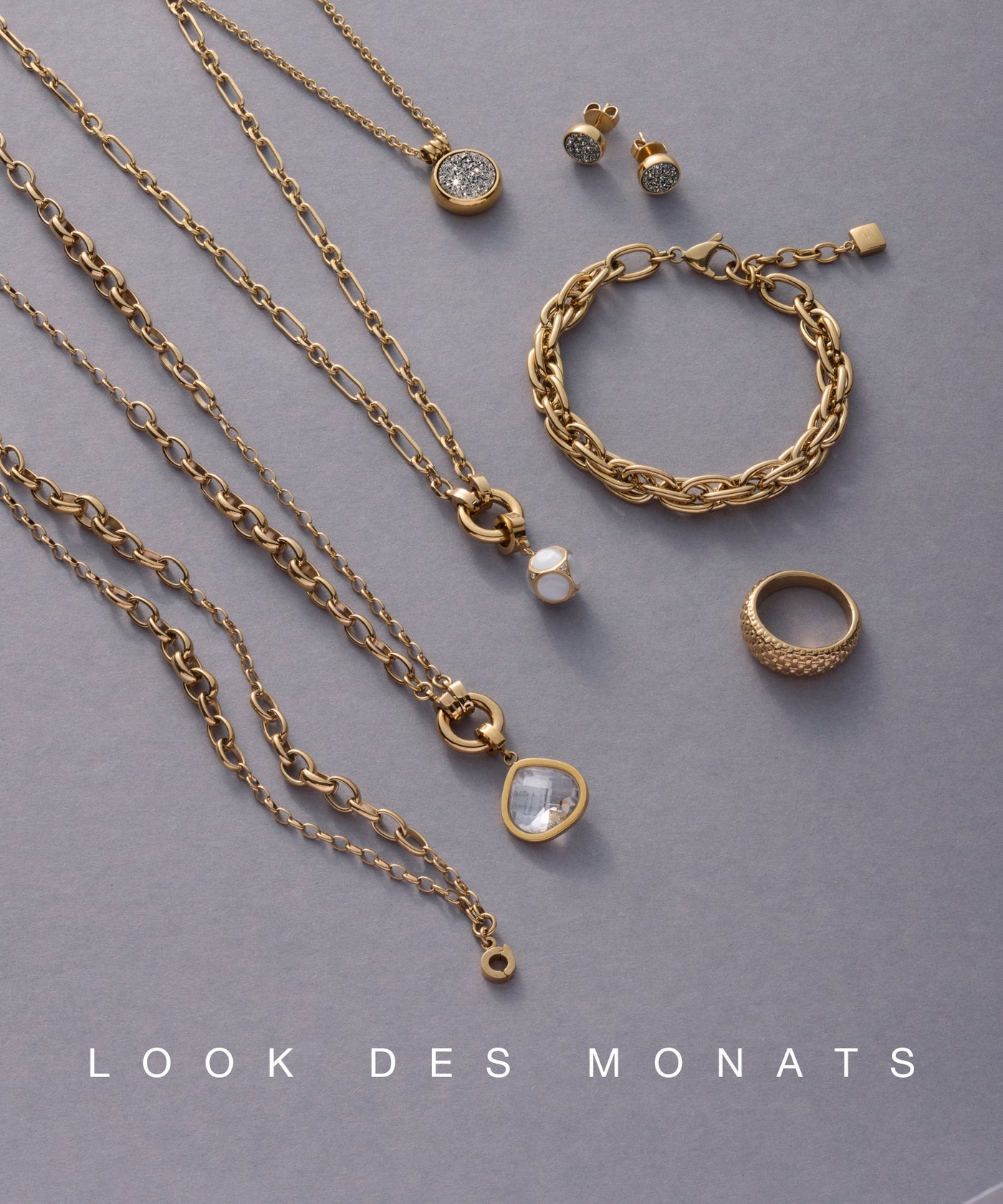 Ein stilvoll arrangiertes Set aus goldfarbenem Schmuck mit Ketten, Ohrsteckern, einem Ring und einem Armband liegt auf grauem Hintergrund – unten steht „LOOK DES MONATS“.