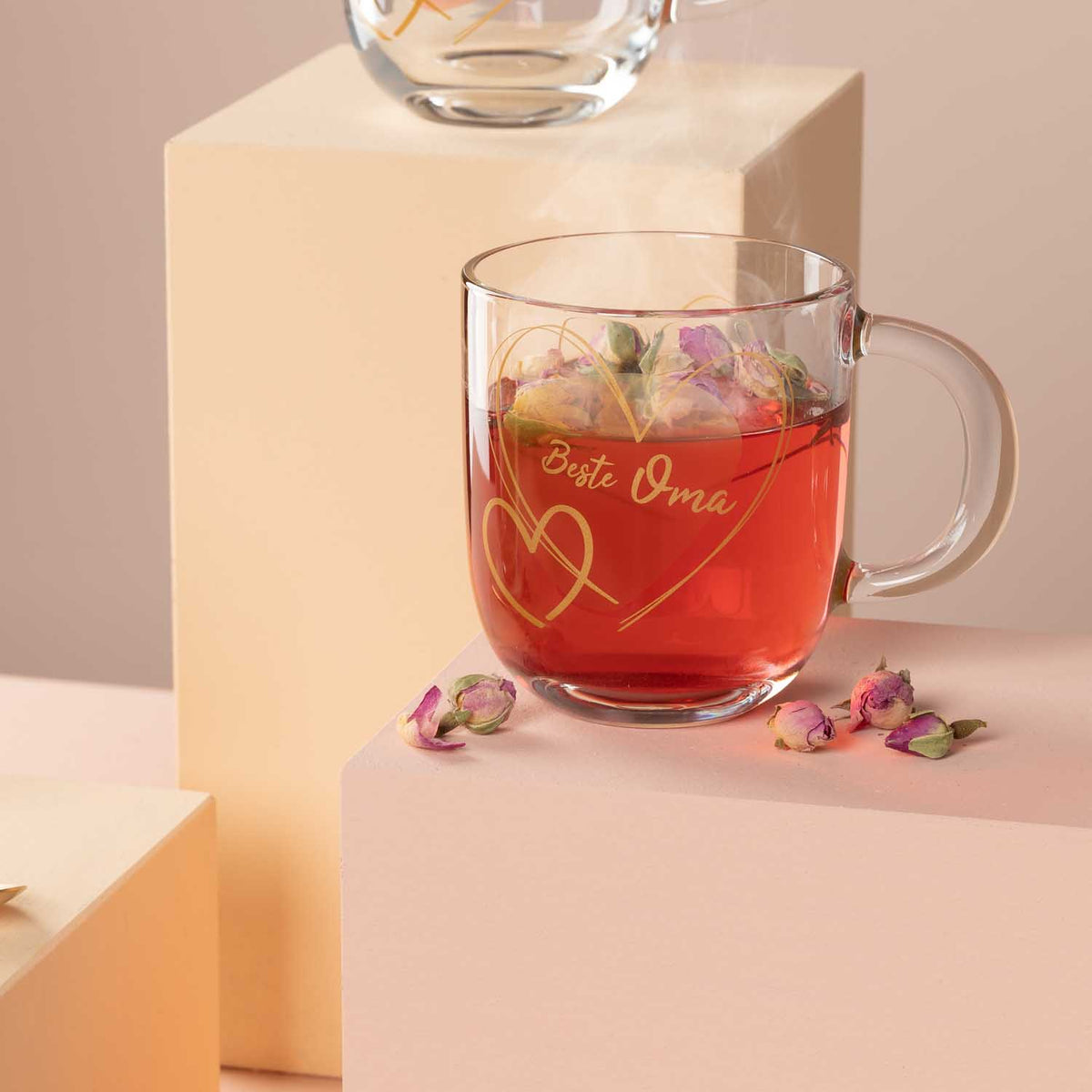 Glastasse EMOZIONE mit rotem Tee, Eiswürfeln und getrockneten Blüten, daneben Rosenblätter auf pastellfarbenem Hintergrund