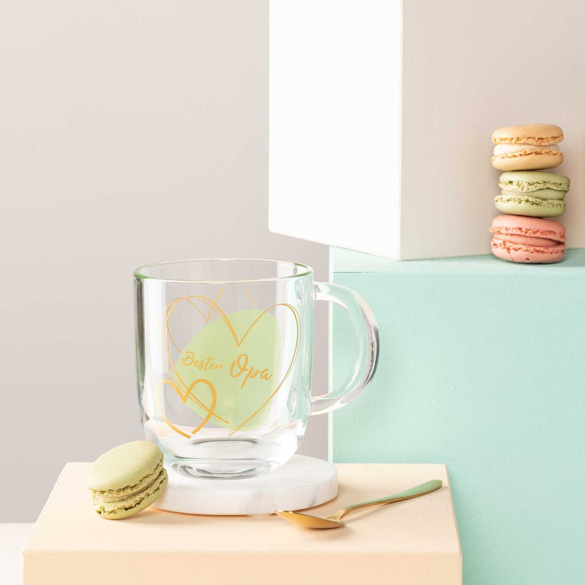 Glastasse mit gelber Gravur "Bester Papa", Macarons und Dekoobjekte auf farbigen Oberflaechen