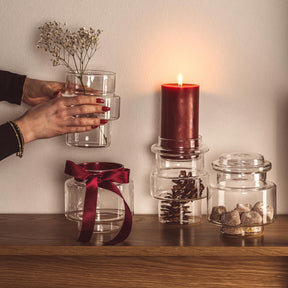 Klare Glsdosen mit Trockenblumen und Pinienzapfen, Kerze und Hand mit Wasser