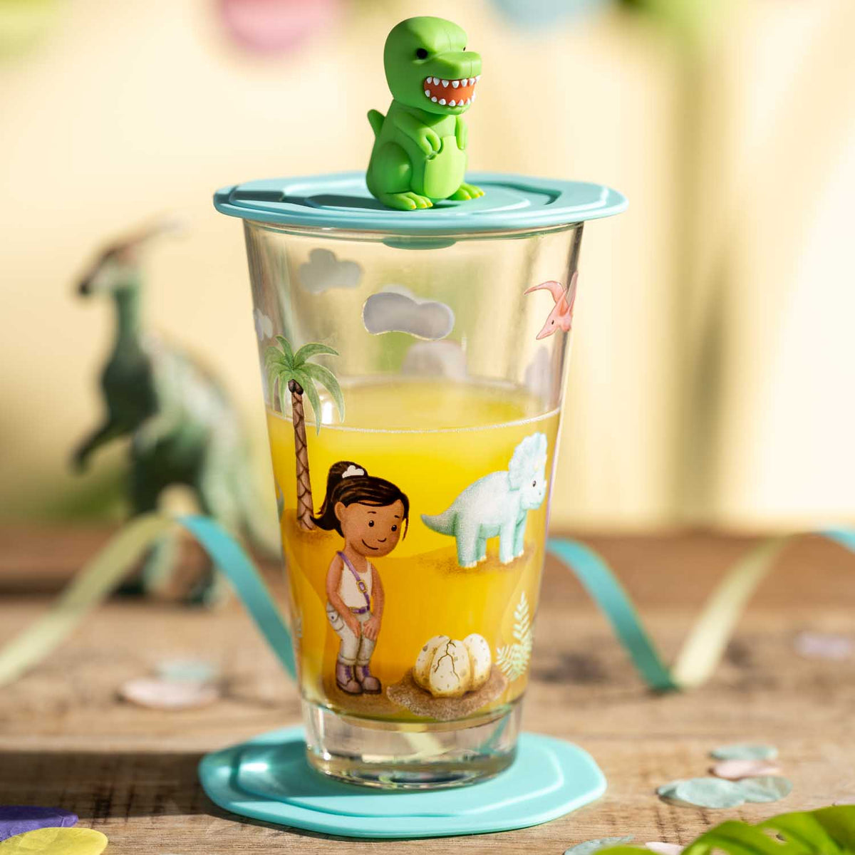 Trinkglas BAMBINI AVVENTURA mit buntem Dino-Motiv und grünem Deckel, daneben Dinosaurier-Spielfiguren