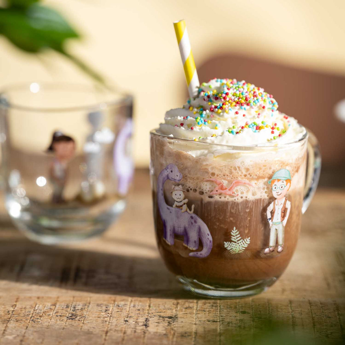 Glastasse BAMBINI AVVENTURA mit bunten Motiven gefüllt mit Kakao, Sahne und Streuseln auf Holztisch