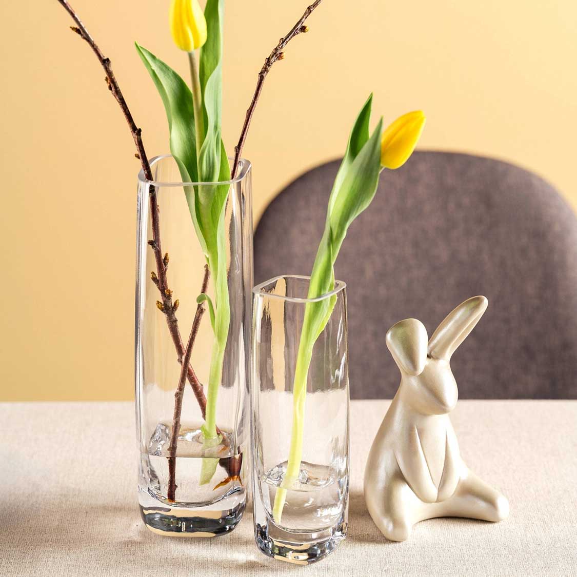 Hase LOLA neben Glasvasen mit gelben Tulpen und Zweigen auf Tisch