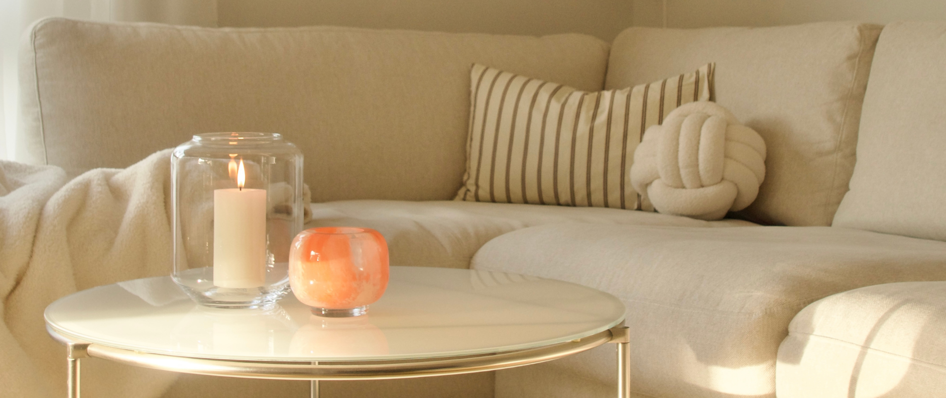 Beiges Sofa mit Kissen, davor ein runder Couchtisch mit brennender Kerze im Glas und kleinem orangefarbenem Windlicht.