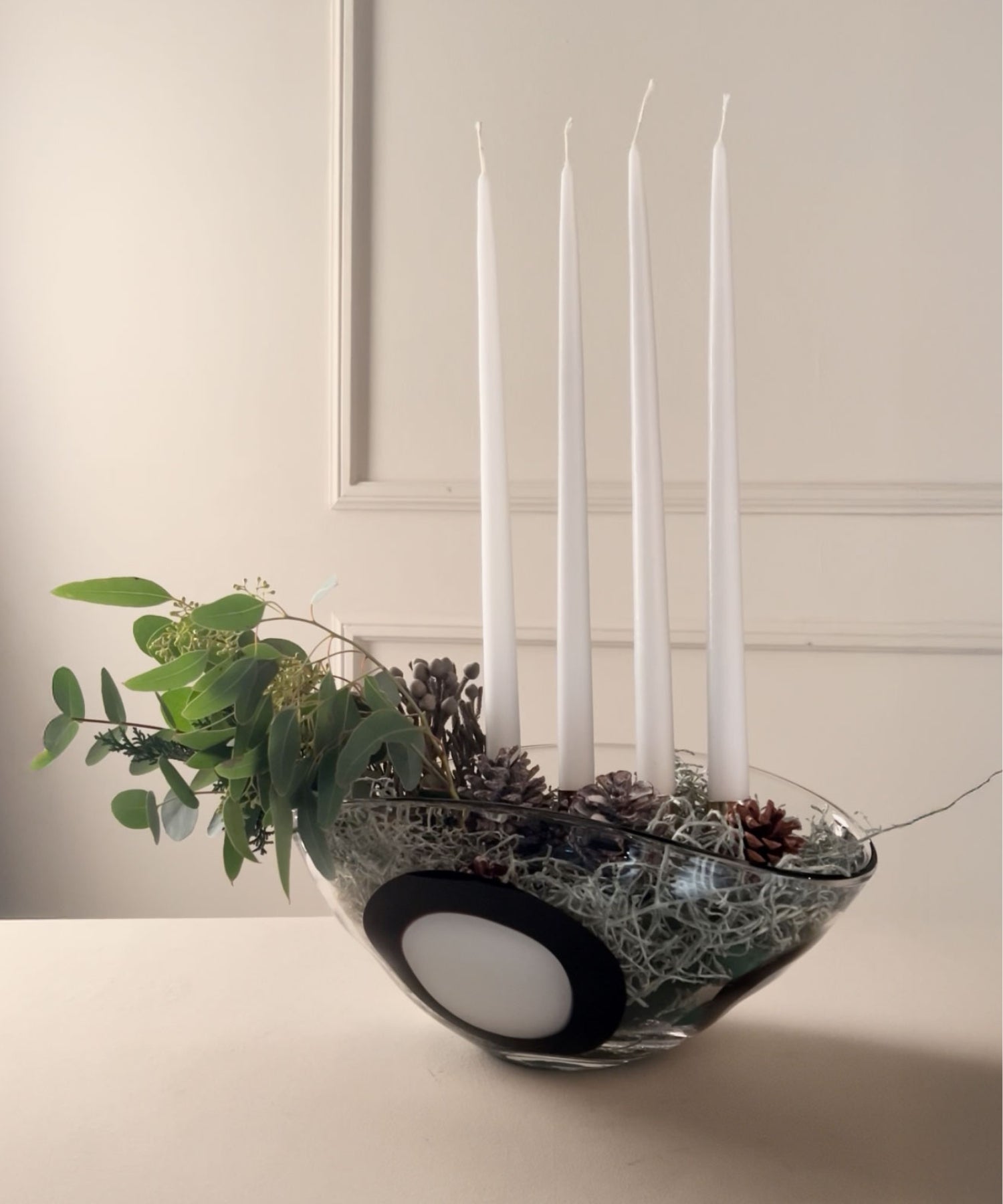 Modernes Adventsgesteck mit vier weißen Stabkerzen in einer Glasschale mit schwarzen Akzenten, geschmückt mit Tannenzapfen, Moos und Eukalyptuszweigen.