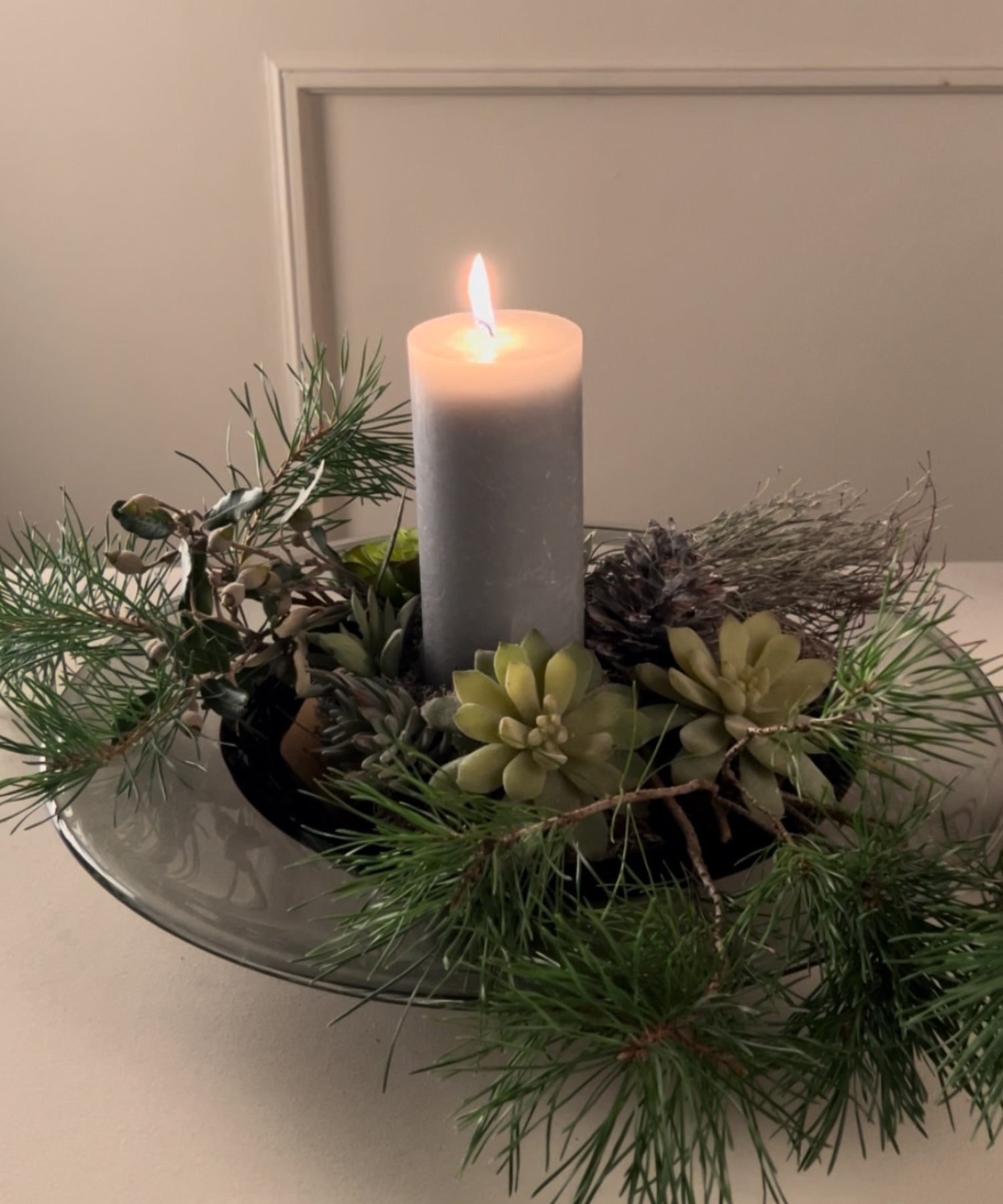 Kleines Adventsgesteck mit einer brennenden grauen Kerze inmitten von Tannenzweigen, Sukkulenten und Zapfen auf einem Glasteller.