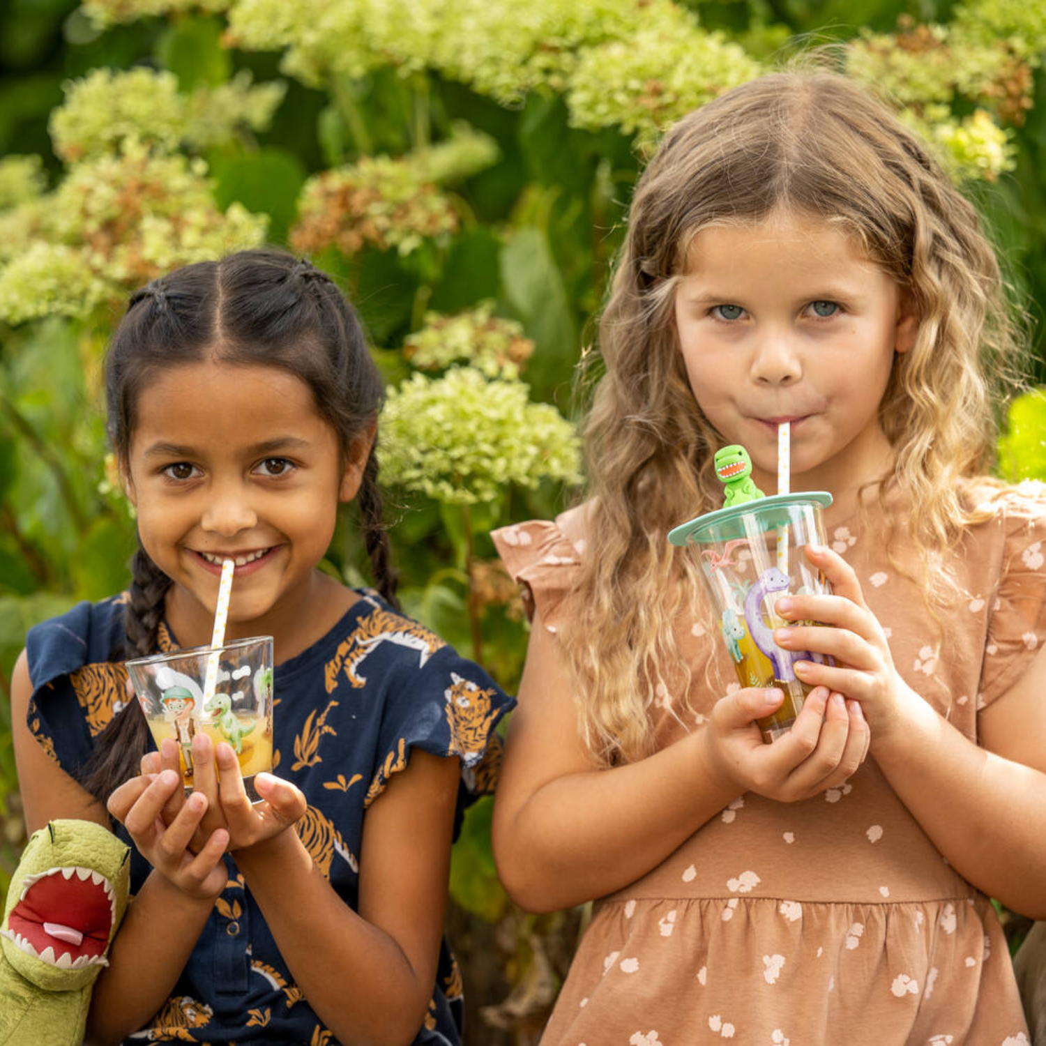 Zwei fröhliche Kinder trinken mit bunten Trinkhalmen aus Gläsern mit Dino-Motiven und genießen den Moment im sommerlichen Garten.