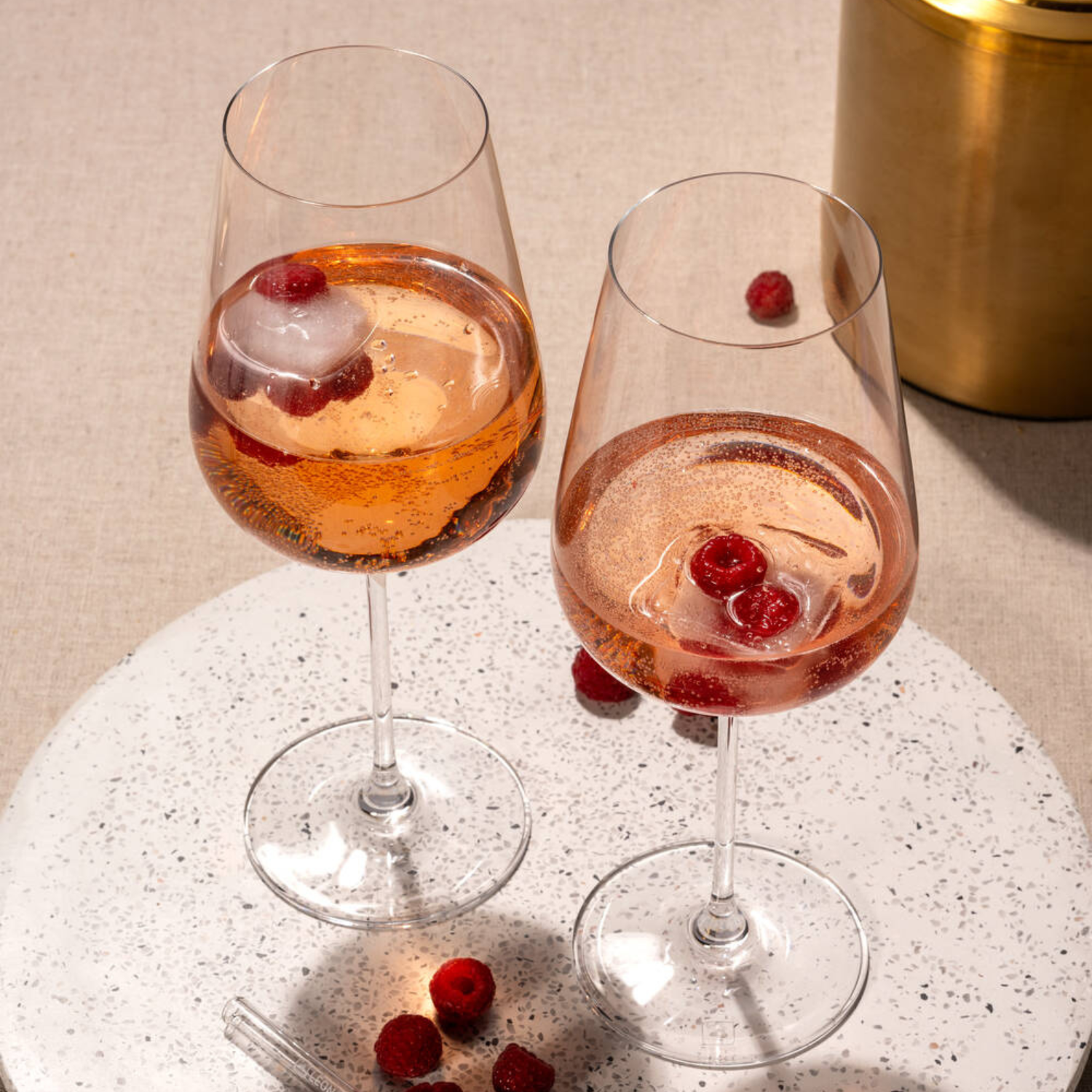 Zwei elegante Weingläser sind mit prickelndem Rosé und gefrorenen Himbeeren gefüllt und stehen dekorativ auf einer hellen Platte.