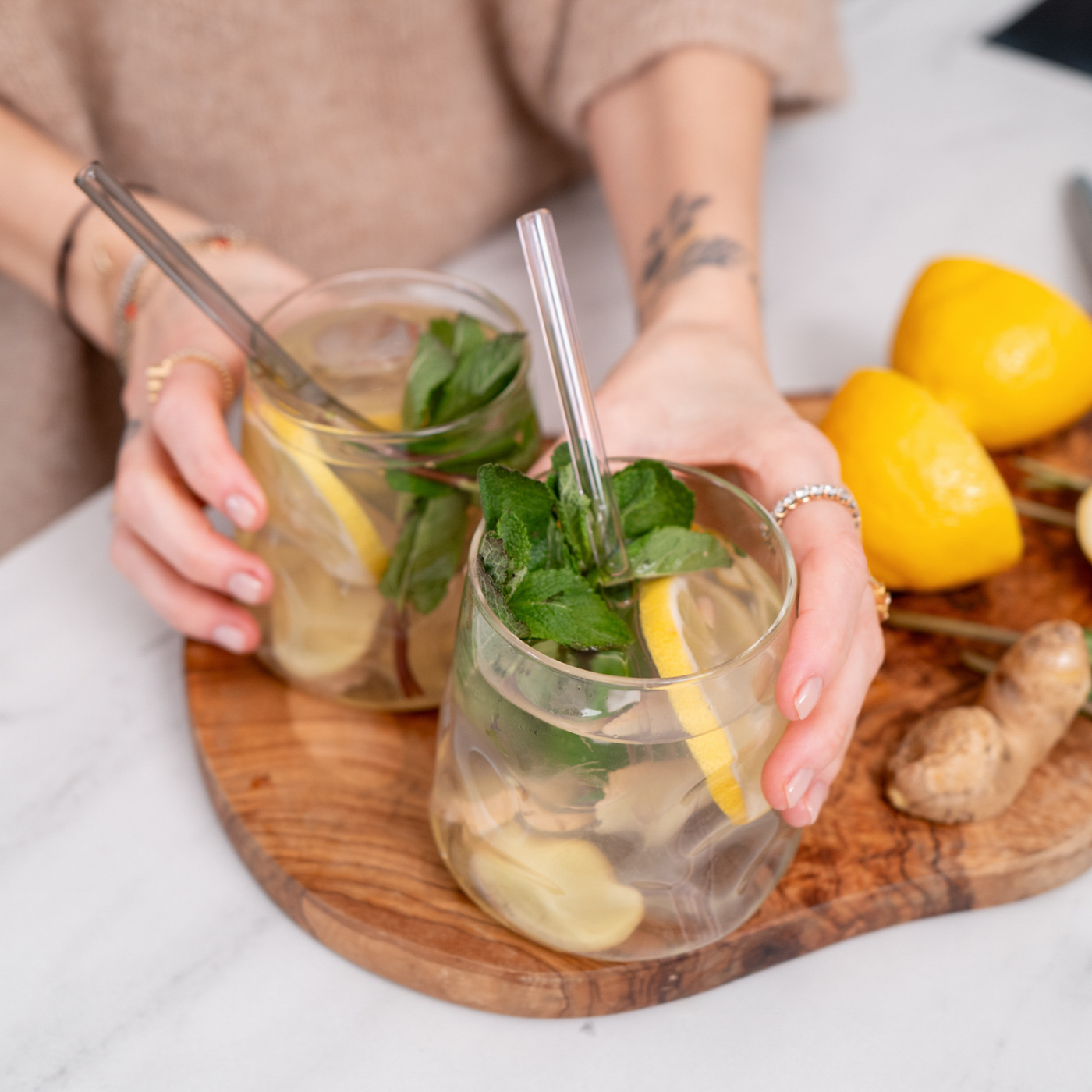 Zwei Glasbecher mit Zitronen-Minz-Wasser und Glasstrohhalmen auf einem Holzbrett, daneben Zitronen und Ingwer.