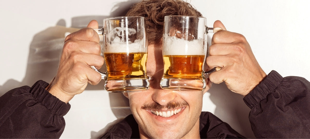 Lachender Mann mit Schnurrbart hält zwei Biergläser mit Bier vor sein Gesicht