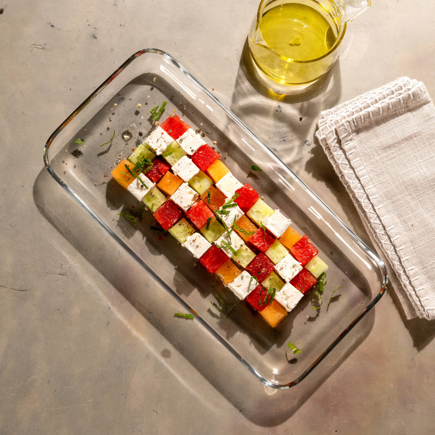 Sommerlicher Würfel-Salat aus Wassermelone, Honigmelone, Gurke und Feta, angerichtet auf einer CIAO Glas-Platte mit Olivenölkanne und Serviette daneben.