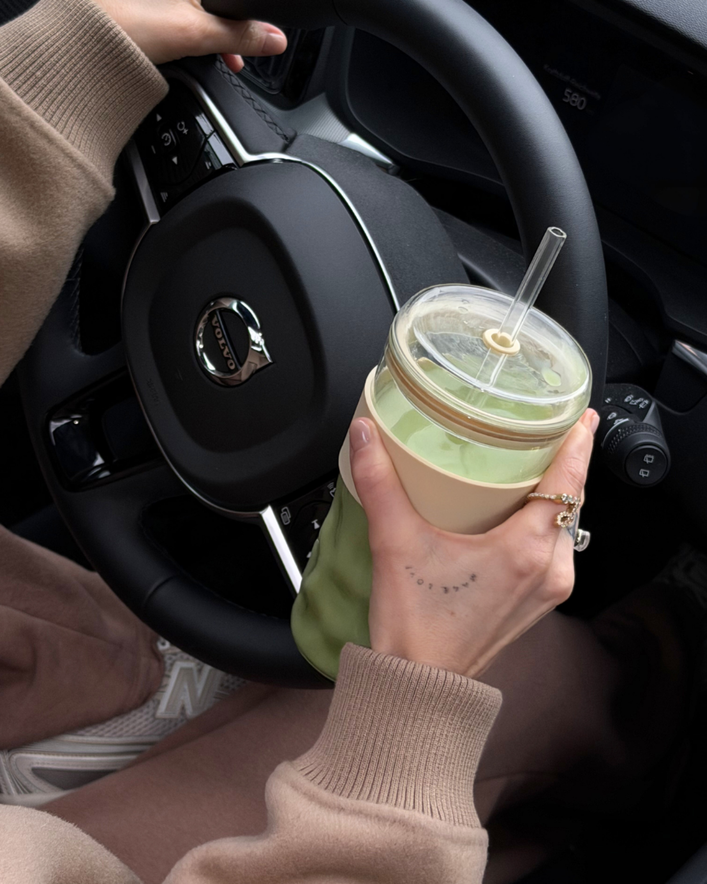 Person hält einen wiederverwendbaren To Go Cup mit grünem Getränk und Glasstrohhalm am Lenkrad eines Autos.