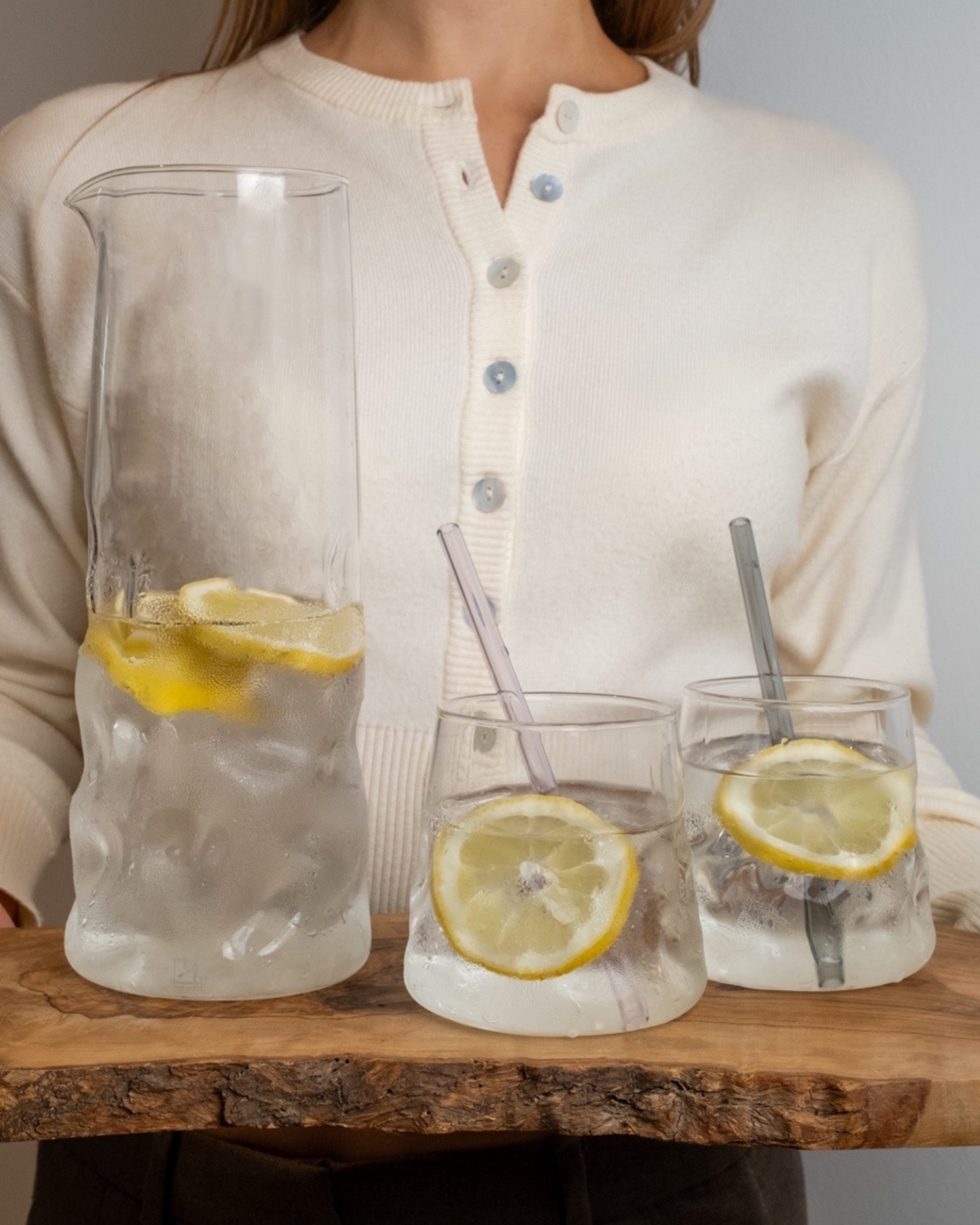 Glaskaraffe und zwei Trinkgläser mit Zitronenscheiben und Wasser auf einem Holzbrett, eine Person steht im Hintergrund.