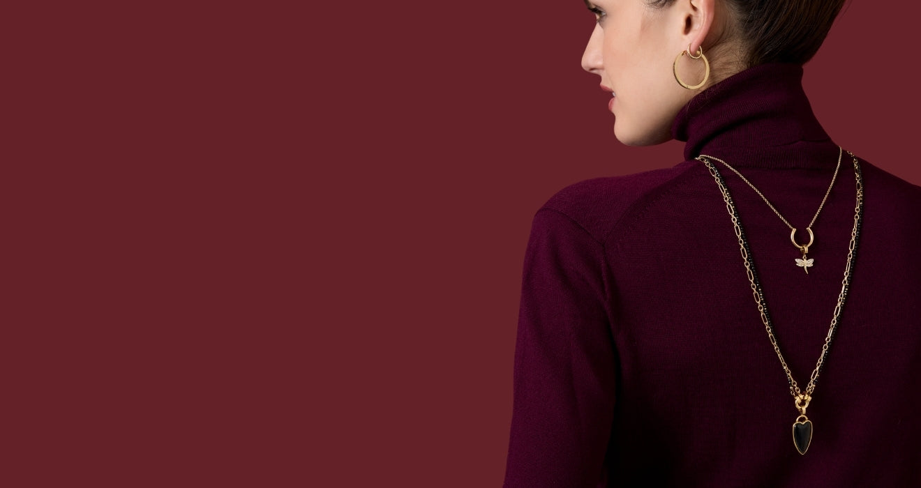 Rückansicht einer Frau mit hochgestecktem Haar, die auffällige, goldene Ketten mit Anhängern über einem weinroten Rollkragenpullover trägt.