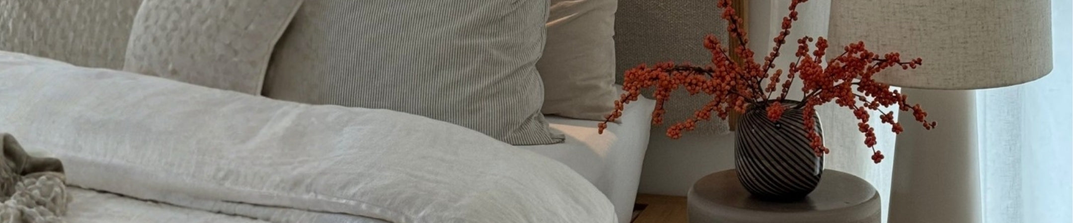 Gemütliches Schlafzimmer mit beigefarbenem Kopfteil, weißen Kissen und roten Beeren in schwarzer Vase auf Nachttisch
