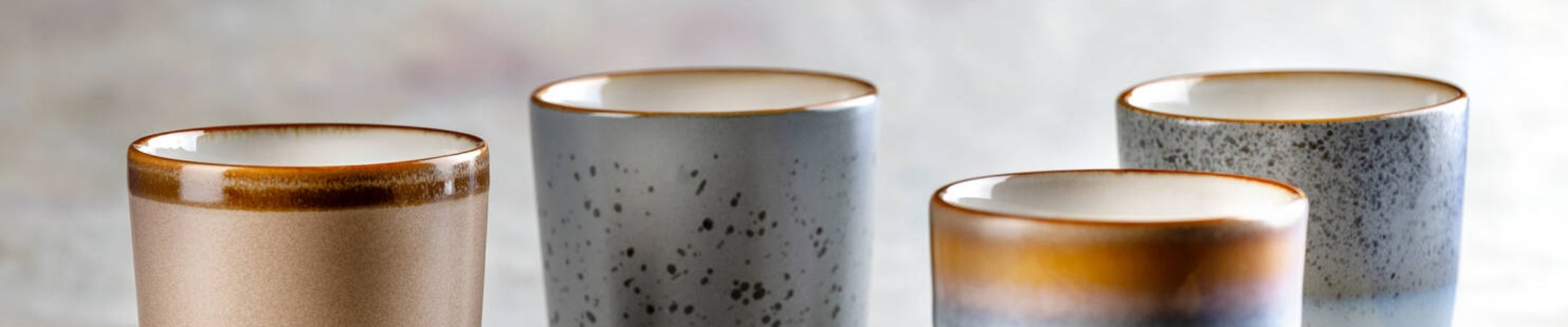 Vier elegante Gläser mit goldenen Rändern in Beige, Grau und Braun-Tönen nebeneinander vor hellem Hintergrund