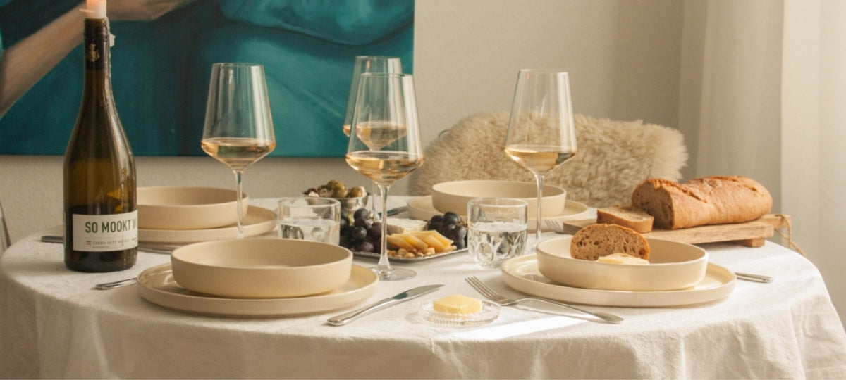 SO MOCKT! Elegantly set table with cream-colored dinnerware, wine glasses, bread, and white wine bottle in warm, minimalist ambiance