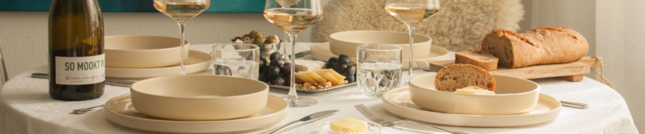 Elegante Tischdeckung mit cremefarbenem Geschirr, Weingläsern, Brot und Flasche SO MOODY auf weißer Tischdecke